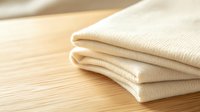 Neatly folded cream cotton fabric napkins or textiles stacked on wooden surface in warm natural lighting, showcasing texture and minimalist style.
