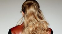 Young woman with long wavy blonde hair wearing brown leather jacket shown from behind against light gray background, modern casual fashion style.