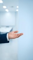 Caucasian male hand in dark business suit and white shirt sleeve gesturing open palm in modern office environment with blurred background. Suitable for business, corporate presentations, and professional concepts.