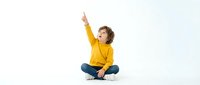 Young boy in yellow sweater sitting cross-legged pointing upward with excited expression on white background for educational and lifestyle content.