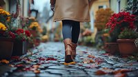 Woman in beige coat and brown leather boots walking on wet cobblestone street with fallen autumn leaves, surrounded by potted chrysanthemums in red and yellow colors.