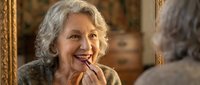 Elderly Caucasian woman with gray curly hair applying lipstick while looking in ornate golden mirror, getting ready for special occasion or daily routine.