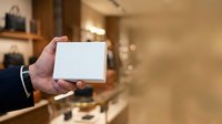 Close-up of male hand holding blank white box in modern retail store interior with warm lighting and blurred shelves background.