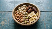 Wooden bowl filled with mixed nuts, dates, and dried banana chips on a rustic blue table, healthy snack concept.