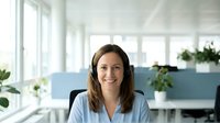 Smiling Caucasian female customer support agent wearing a headset sits at a bright modern office, ready to assist clients.