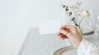 Woman's hand holding blank white card at elegant table setting with white flowers and dinnerware for wedding, event planning, invitation, or stationery mockup.