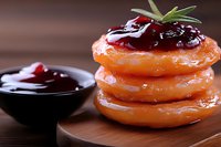 Glazed donuts stacked with cherry jam topping and fresh rosemary garnish on wooden board, dark moody background with side bowl of red fruit preserve.