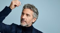 Mature man with gray hair and beard wearing navy blazer and black turtleneck, touching head against blue background. Suitable for business, corporate, lifestyle, and professional portraits.