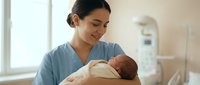 Young Hispanic female nurse in blue scrubs gently holding newborn baby in hospital maternity ward with soft natural lighting from window.