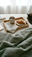 Cozy breakfast in bed with croissant, tea cup on wooden tray, stay home napkin, green blanket, and cat resting nearby in soft morning light.
