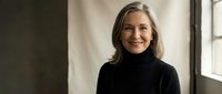 Confident mature woman with gray hair smiling in black turtleneck sweater standing by window in modern office or home interior for business portrait.