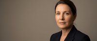 Middle-aged woman with dark hair and fair complexion wearing dark blazer against neutral background, suitable for corporate, business, and professional headshot uses.