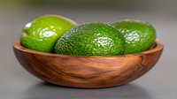 Fresh ripe whole avocados in wooden bowl against neutral background, selective focus on textured green skin. Food photography for healthy eating concept.
