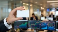 Close-up of businessman's hand holding blank white business card in modern office with stock market displays and colleagues in background.