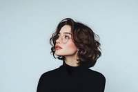 Young Asian woman with short wavy brown hair wearing glasses and black turtleneck sweater poses against light blue background in elegant minimal portrait.