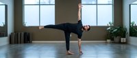Young man with dark hair and light skin tone performing a yoga warrior pose in a bright studio with large windows, potted plants, and wooden flooring for fitness and wellness content.