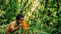 Young boy with dark skin tone and curly hair wearing orange shirt smelling bright yellow-orange tropical flower in lush green jungle foliage environment. Nature exploration and botanical discovery.