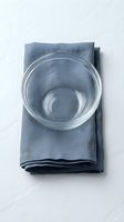 Empty glass bowl on folded blue linen napkin against white marble surface, overhead view. Suitable for food styling, recipe blogs, kitchen product photography.