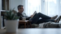 Middle-aged Caucasian man with glasses reading book while relaxing on couch at home. Comfortable indoor lifestyle scene for leisure and education concepts.