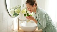 Woman with medium skin tone in casual green shirt washing face over white ceramic sink in bright bathroom with fresh flowers and mirror reflection.