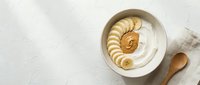 Creamy yogurt bowl topped with sliced bananas and peanut butter swirl, served with wooden spoon on white textured surface for breakfast and healthy eating content.