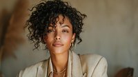 Young African American woman with curly hair wearing beige blazer and gold necklace, looking confidently at camera against neutral background.