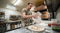 Male baker with dark hair and beard, wearing apron, dusting flour onto pizza dough in professional kitchen bakery setting with industrial equipment and ingredients visible.