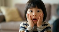 Young Asian girl with hands on cheeks showing surprised expression wearing striped sweater in cozy living room setting for childhood emotion concepts.