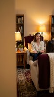 Young Caucasian woman relaxing in cozy living room with warm lighting, bookshelves, and comfortable armchair for home lifestyle content.