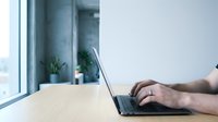 Professional businessman typing on laptop computer at modern office desk near window with natural light for remote work and business productivity.