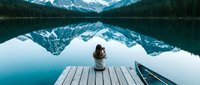 Solitary woman sitting on wooden dock gazing at snow-capped mountain reflected in calm turquoise alpine lake surrounded by evergreen forest.