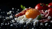 Raw egg yolk with prosciutto, fresh tomatoes and crumbled feta cheese on dark background with scattered sea salt and herbs, dramatic food photography.