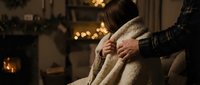 Cozy winter evening at home, man wrapping woman in warm blanket by fireplace with Christmas lights glowing in background.