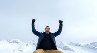 Young man with arms raised in celebration on snowy mountain peak, expressing joy and achievement in winter landscape setting.