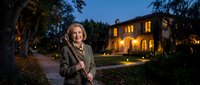 Confident mature Caucasian businesswoman in elegant coat standing outdoors at twilight with illuminated luxury home in background for real estate marketing.