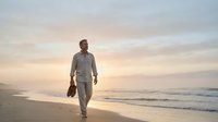 Middle-aged man in casual business attire walking barefoot along sandy beach at sunset, carrying shoes in hand, serene coastal landscape with calm waves and golden sky.