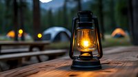 Glowing oil lamp on wooden table at dusk with camping tents in background. Atmospheric outdoor scene captures wilderness adventure and night camping experience.