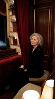 Elegant senior woman with curly gray hair in black velvet dress sitting in ornate theater box with red curtains and golden details for luxury entertainment concept.