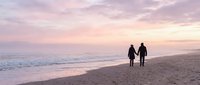 Romantic couple silhouettes walking hand in hand on sandy beach at sunset with pink purple sky and ocean waves for love and travel concepts.