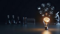 Glowing lightbulb with brain filament emitting business icons on dark background symbolizing innovation and creative thinking in corporate environment.