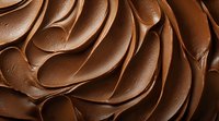 Creamy chocolate frosting swirls in rich brown waves creating luxurious texture pattern, macro close up view shows smooth velvety surface with delicate peaks and curves.