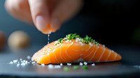Fresh raw salmon fillet garnished with sea salt flakes and chopped chives on dark slate background, with hand sprinkling seasoning from above.