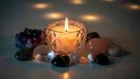 Glowing candle in crystal holder surrounded by colorful gemstones and healing crystals creating peaceful meditation atmosphere with warm ambient lighting.