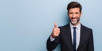 Confident businessman in formal black suit showing thumbs up against light blue background, expressing approval and satisfaction.