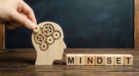Hand placing wooden gear puzzle piece into human head silhouette next to mindset letter blocks on desk with chalkboard background for business growth.