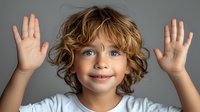 Cheerful Caucasian child with curly blonde hair wearing white t-shirt raises hands up with excited expression against gray background, showing joy and happiness.