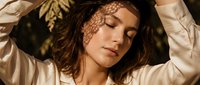 Woman with long brown hair wearing white shirt, eyes closed, relaxed pose with arms raised behind head, natural sunlight casting leaf shadows on face and body, peaceful expression, suitable for wellness and lifestyle content.