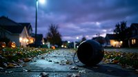 Black soccer ball on wet asphalt street at dusk with glowing streetlights and residential houses creating atmospheric evening sports scene for recreation themes.