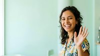 Young woman with dark wavy hair and South Asian ethnicity wearing a colorful patterned shirt, waving and smiling warmly in a bright office environment with green-tinted walls.