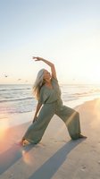Young blonde woman practicing yoga warrior pose on sandy beach at golden hour sunrise with ocean waves and seagulls flying in peaceful morning light.
